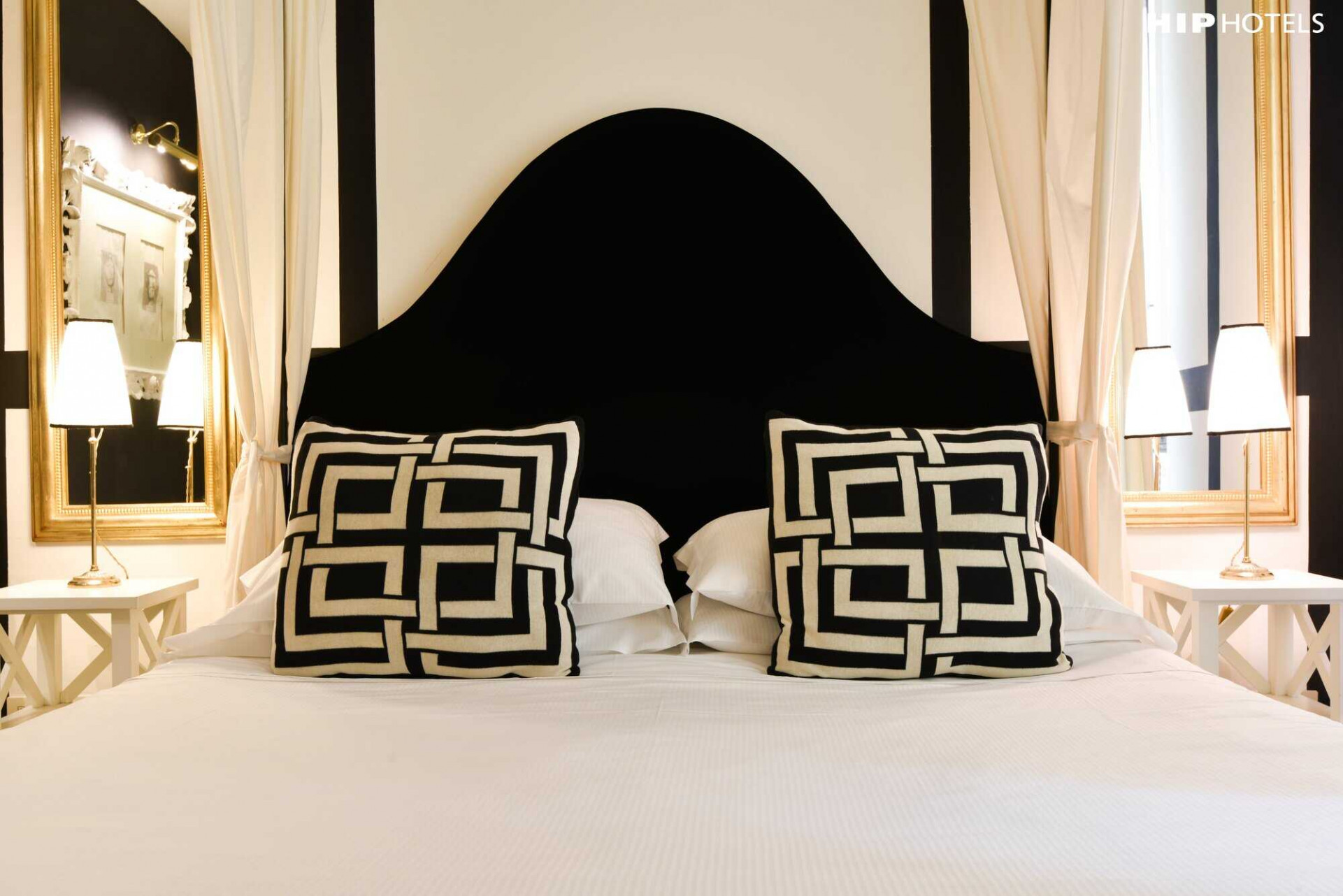 Cama del Hotel Cellai con cojines y decoración en tonos negros, blancos, y dorados. 